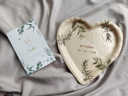 Witte schaal in hartvorm met groene bladeren en een tekst, naast een kaart met bloemen.