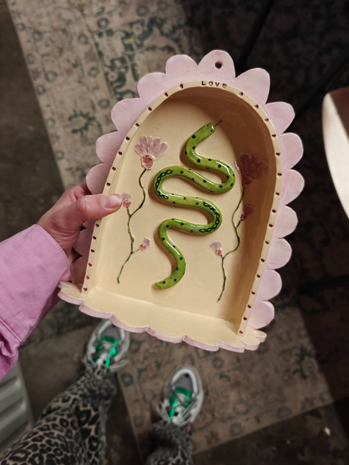 Hand houdt een decoratieve schaal met een groene slang en bloemen. Sneakers zichtbaar.