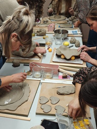 Groep mensen werkt aan kleiprojecten op een tafel, met materialen verspreid rondom.
