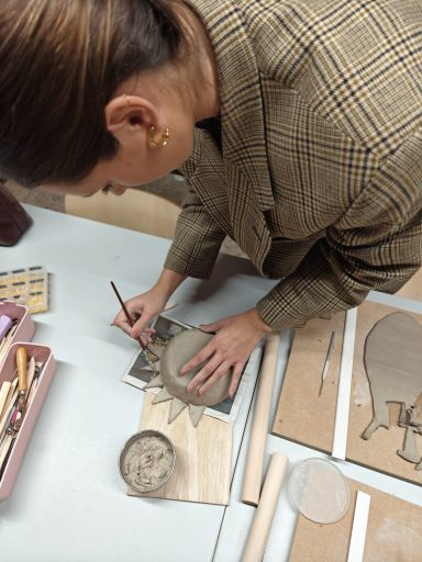 Een persoon werkt met klei en gereedschap op een tafel met materialen en ontwerpen.