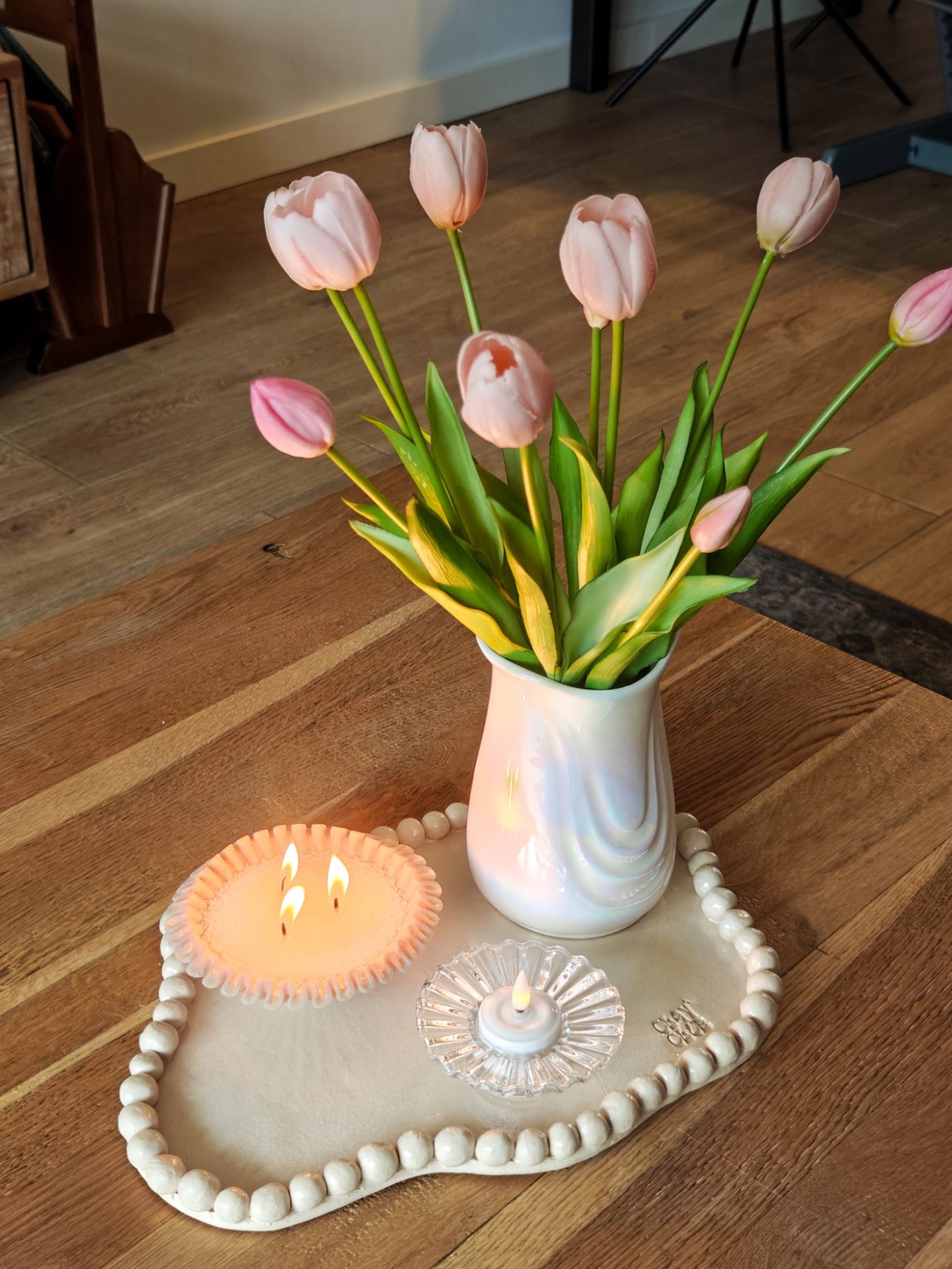 Een vaas met roze tulpen, een kaars en een decoratief bord op een houten vloer.