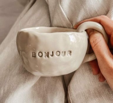 Witte mok met het woord "BONJOUR" in reliëf en een hand die de mok vasthoudt.