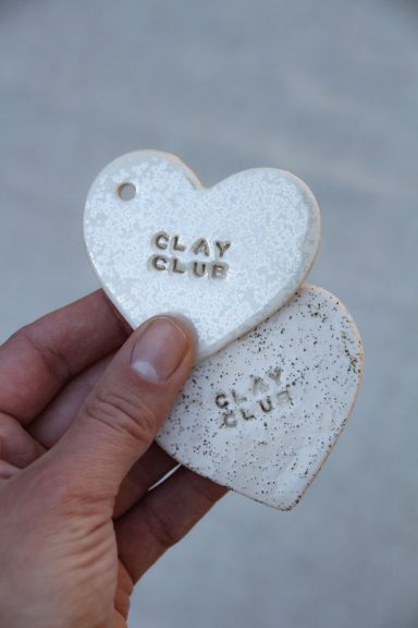 Twee licht gekleurde hartvormige tags met de tekst "CLAY CLUB" erin geperst.
